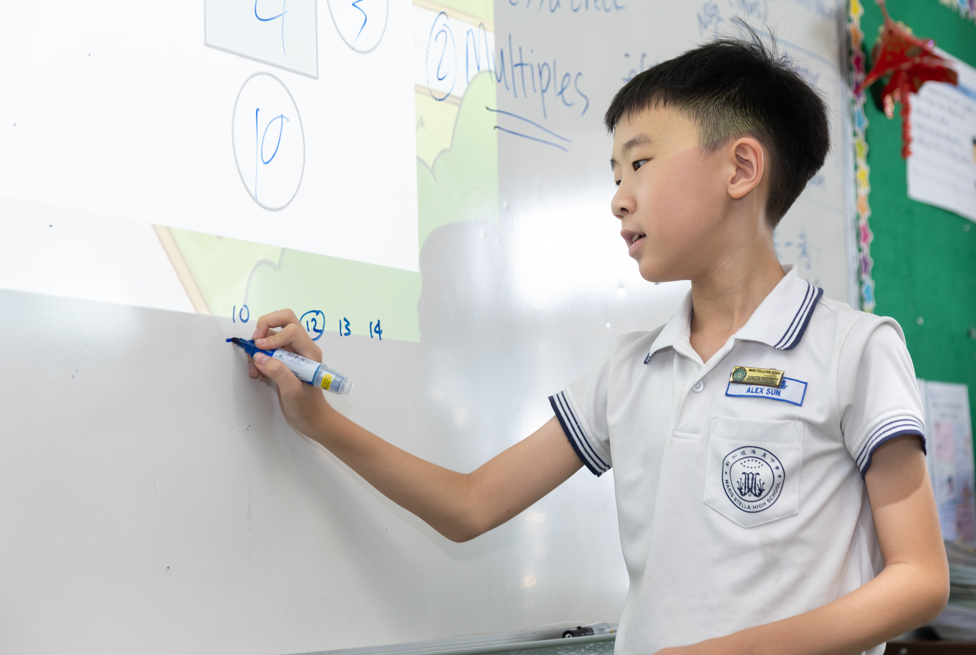 Three young students in white uniforms sit together at a table, smiling and engaged in a classroom activity.